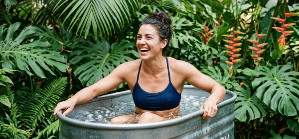 Woman laughing while sitting in a metal cold plunge tub labeled Recovery, surrounded by tropical plants