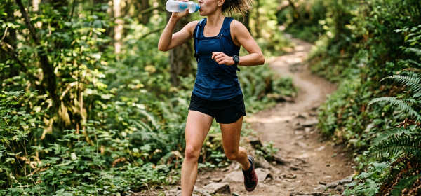 Female endurance athlete drinking from a hydration bottle while trail running through a forest
